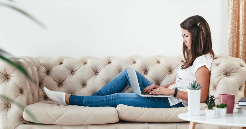 A woman lounges on her couch, working from home on her laptop. 