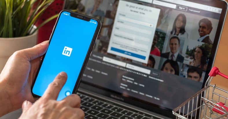 User holding a mobile phone with the LinkedIn icon on the phone screen. There is a laptop in the background with the login screen for LinkedIn 