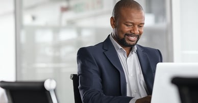 A man in a suit smiles and works on a laptop.