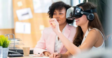 Two women sit a desk, with one woman wearing a VR headset.